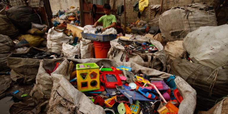 A child sorts through electronic and plastic waste in a landfill, surrounded by large bags of discarded items.