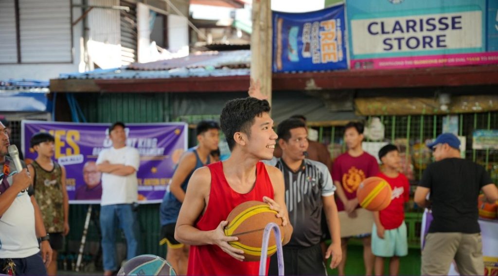 A young basketball player in a red jersey holds a ball while smiling during the Yes Na All 3x3 Basketball League, with spectators and a referee in the background.