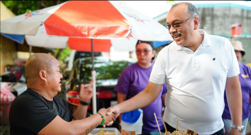 Allan Reyes warmly shaking hands with a local vendor at an outdoor market.